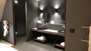 A modern bathroom with a dark tiled wall, a large mirror, and a sink with a black basin. There is a towel rack with towels and a small wooden shelf with a towel and a pair of shoes.