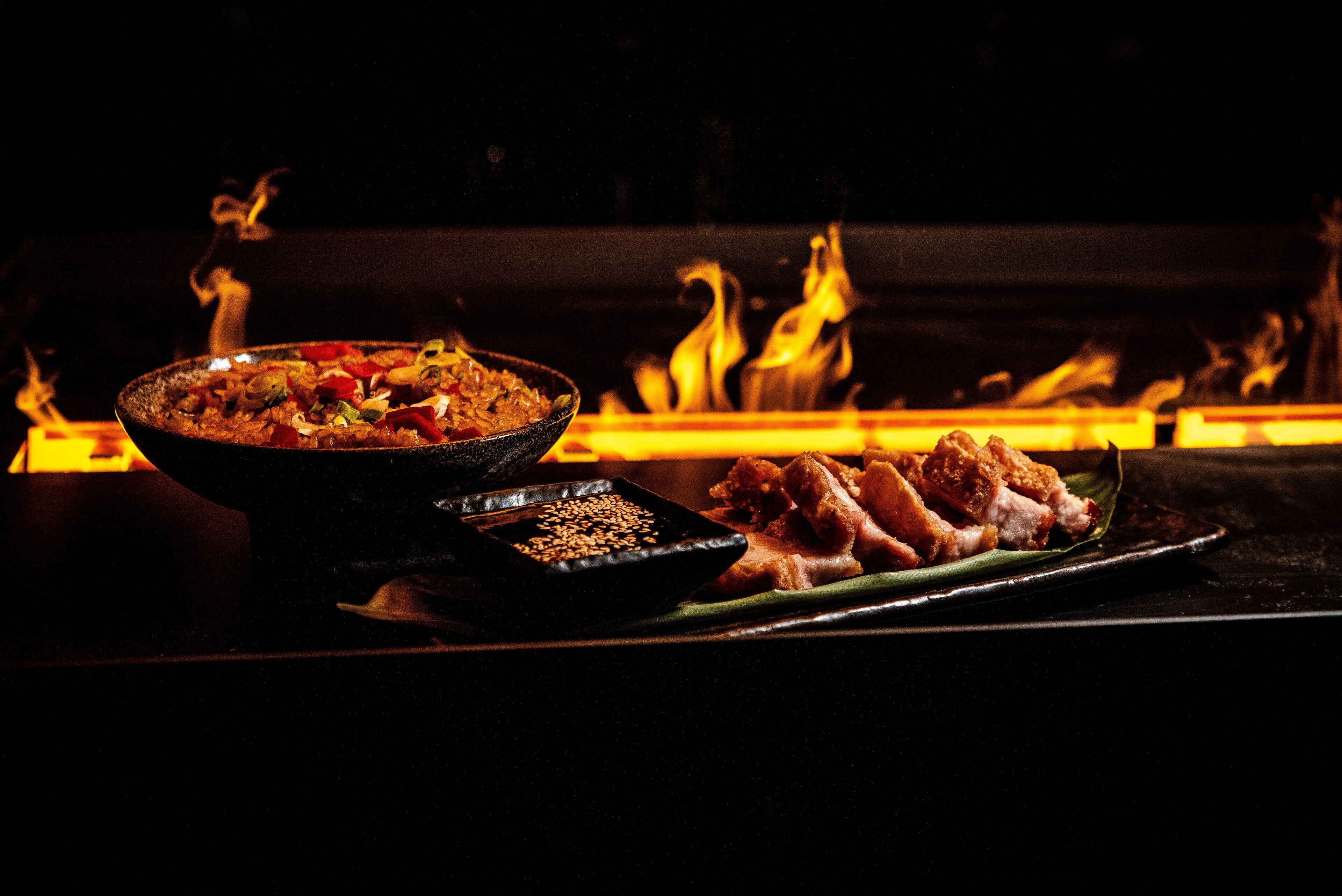A table with a bowl of food and a plate of meat, set against a background of flames.