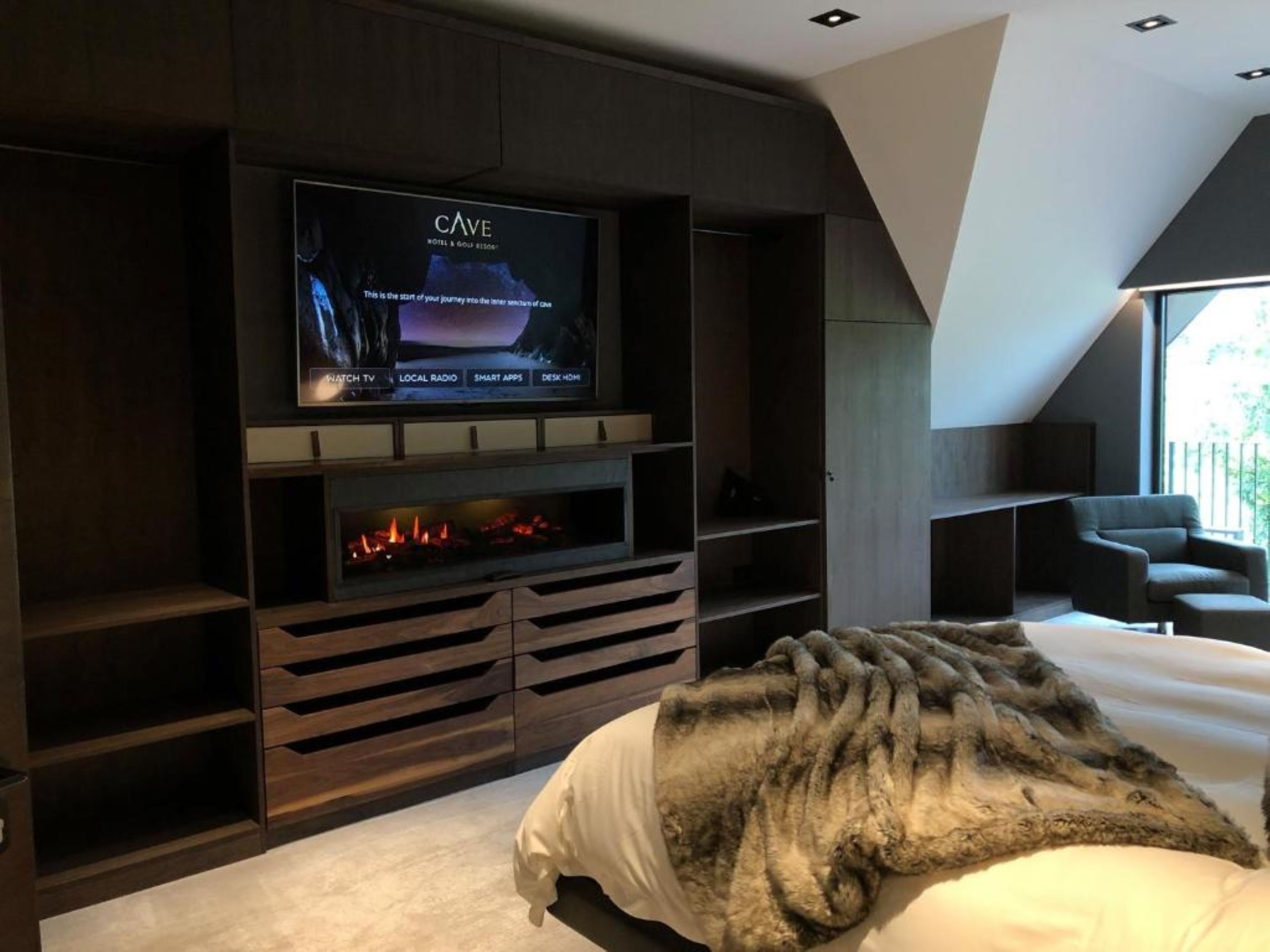A modern bedroom with a TV mounted on the wall, a fireplace, and a cozy bed with a blanket.