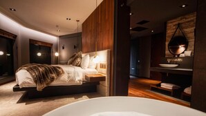 A modern bedroom with a large bed, a bedside table, a wall-mounted mirror, and a bathtub. - Cave Hotel & Golf Resort (Faversham)