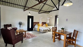 A room with a bed, a chair, a table, a fan, and a potted plant.