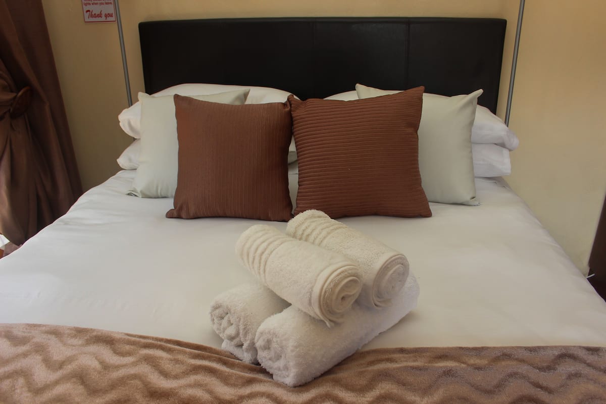 A neatly made bed with white and brown pillows, a brown blanket, and two rolled towels.
