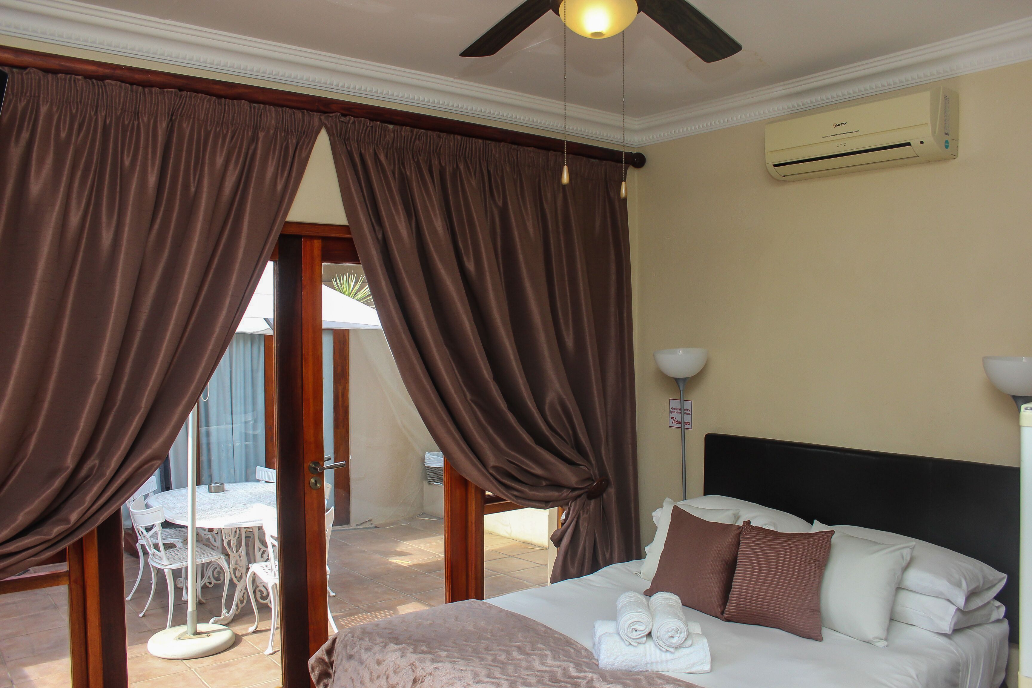 Habitación de hotel con cama, cortinas, ventilador de techo, aire acondicionado, una mesa y sillas afuera, y vistas al exterior.