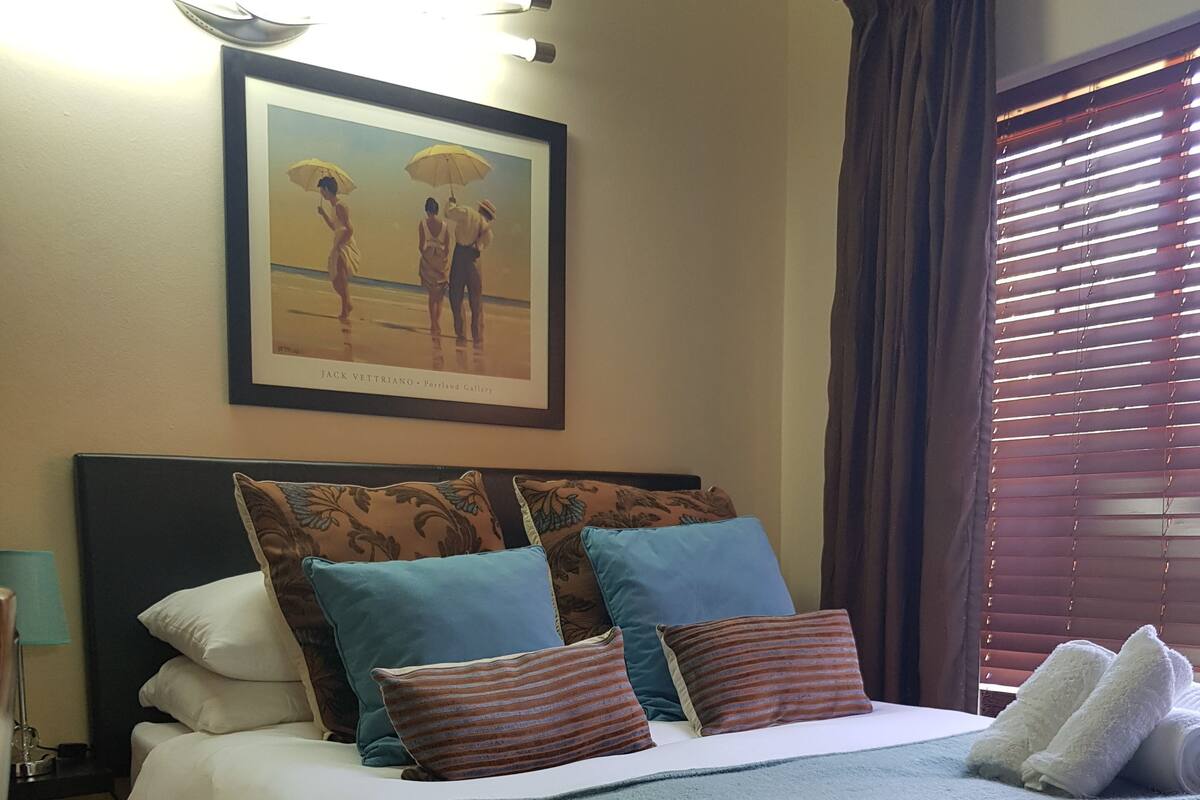 A neatly made bed with a blue throw blanket and pillows, a framed painting above it, and a window with blinds.