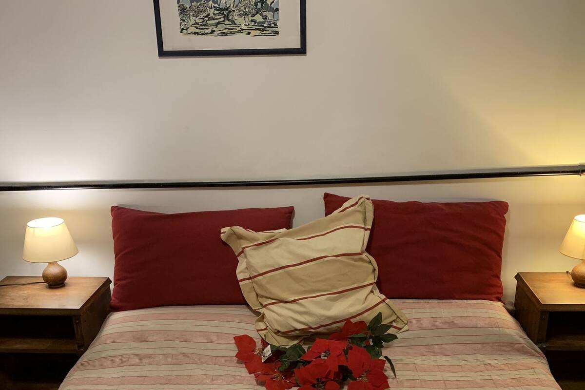 Una cama bien hecha con almohadas rojas y beige, un edredón a rayas y un pequeño ramo de flores rojas. Hay dos lámparas de noche de madera en mesitas auxiliares.
