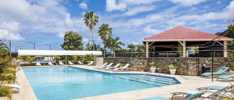 A swimming pool with lounge chairs and a covered area, surrounded by palm trees and a stone wall.