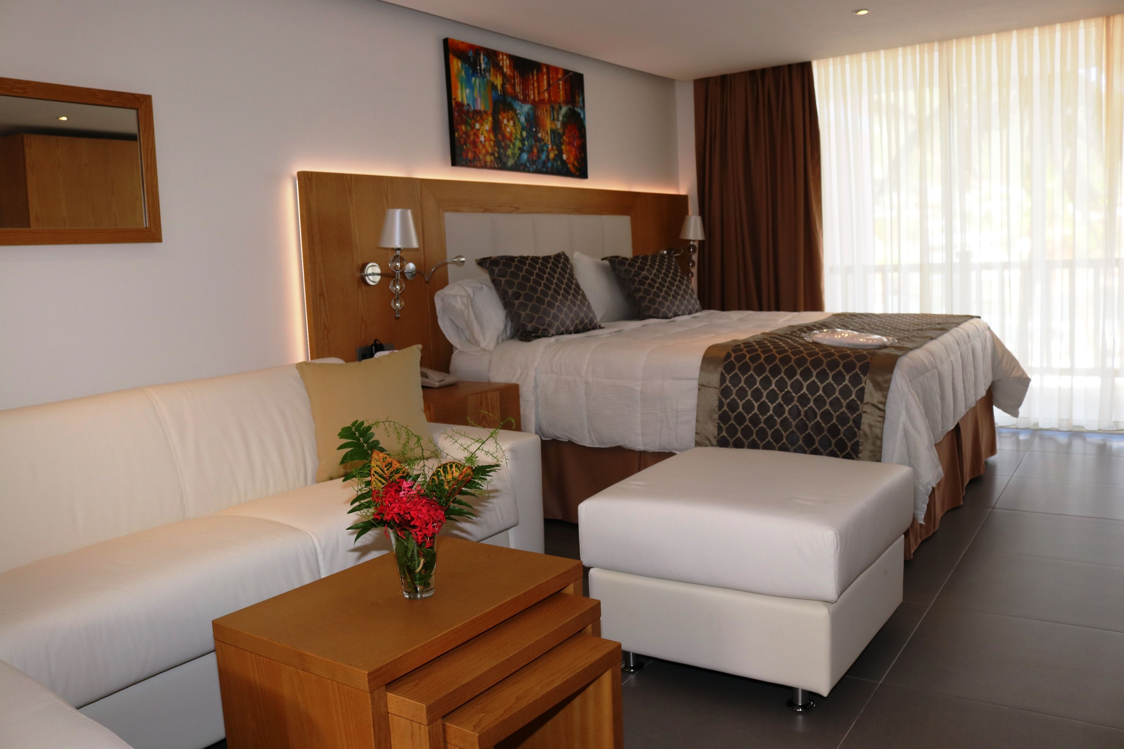 A modern hotel room with a large bed, a sofa, a wooden side table, and a vase of flowers.