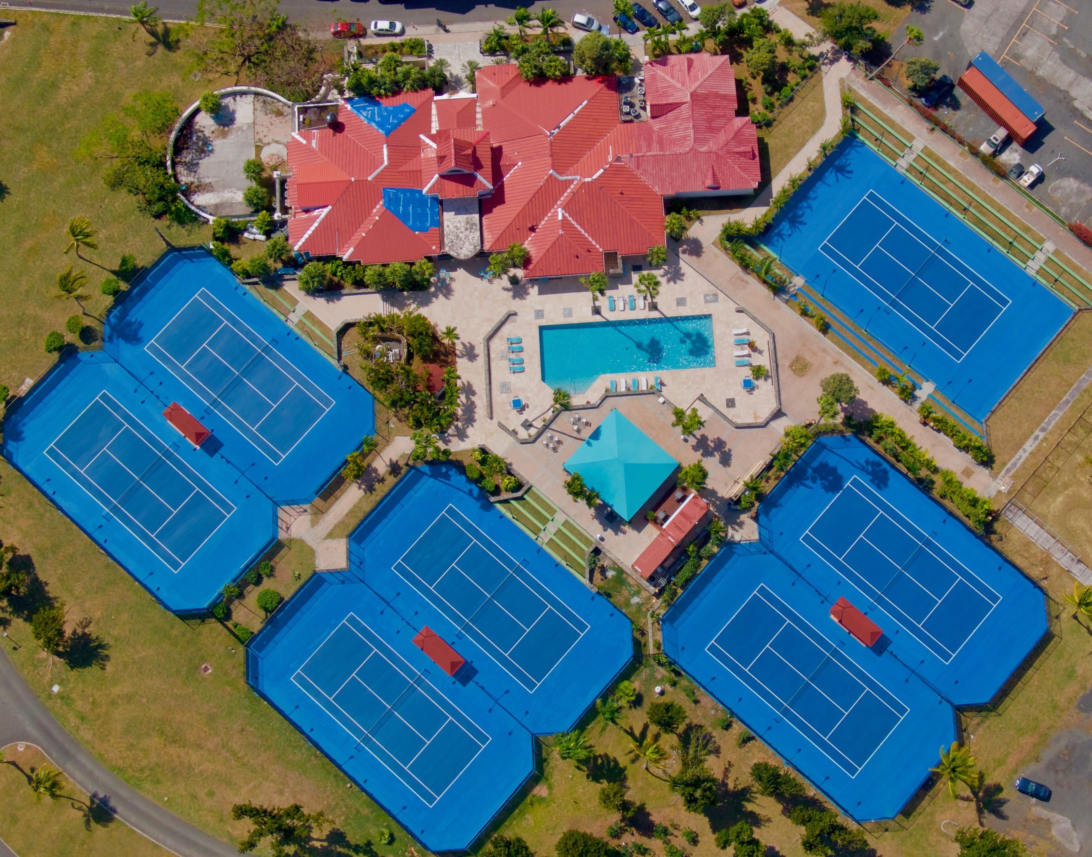 Oversigtsbillede af et sportskompleks med flere tennisbaner, en swimmingpool og en central bygning med rødt tag.
