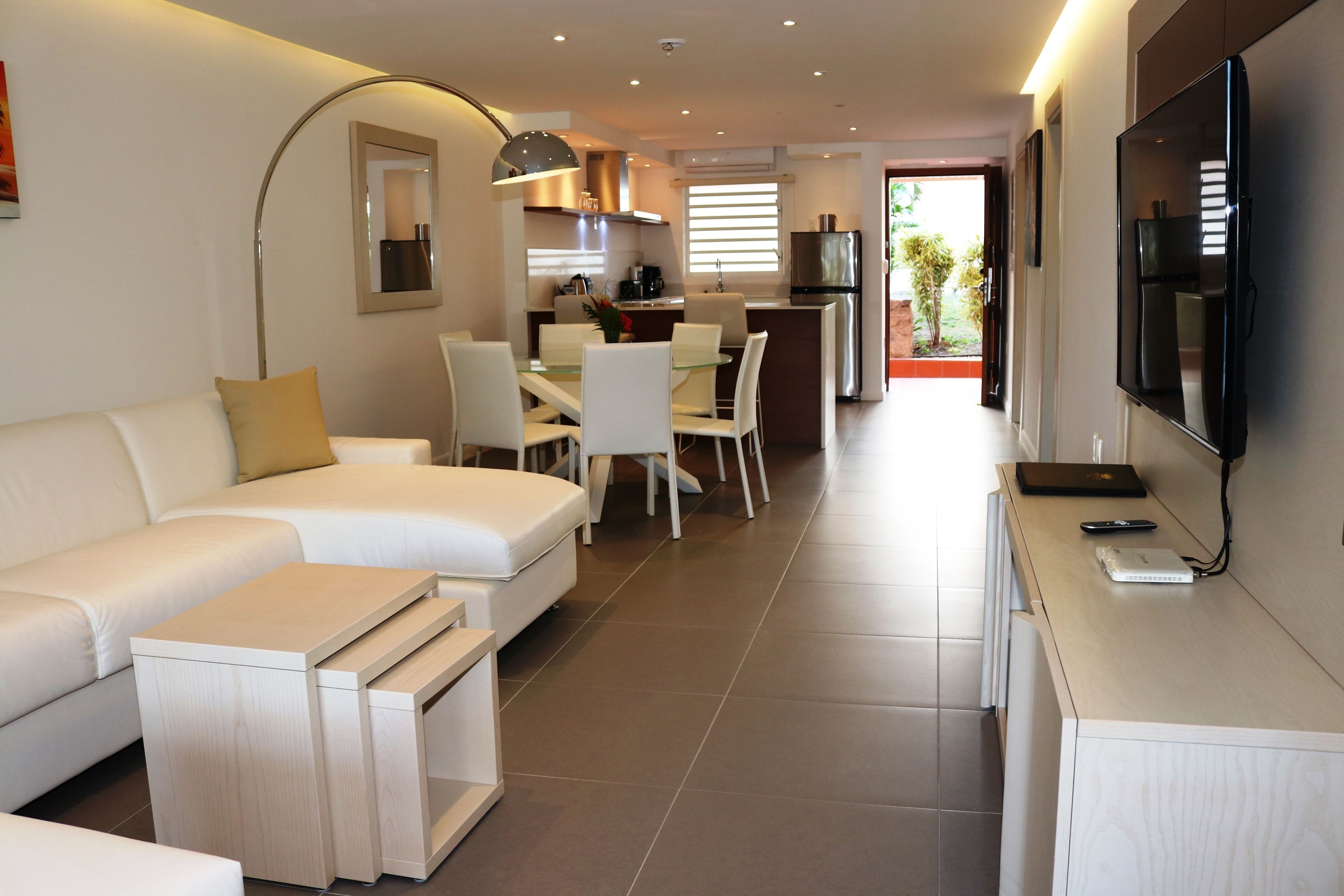 A modern living room with a sofa, coffee table, and TV. There is a dining area with chairs and a table. The room has a kitchen in the background with a refrigerator and stove.