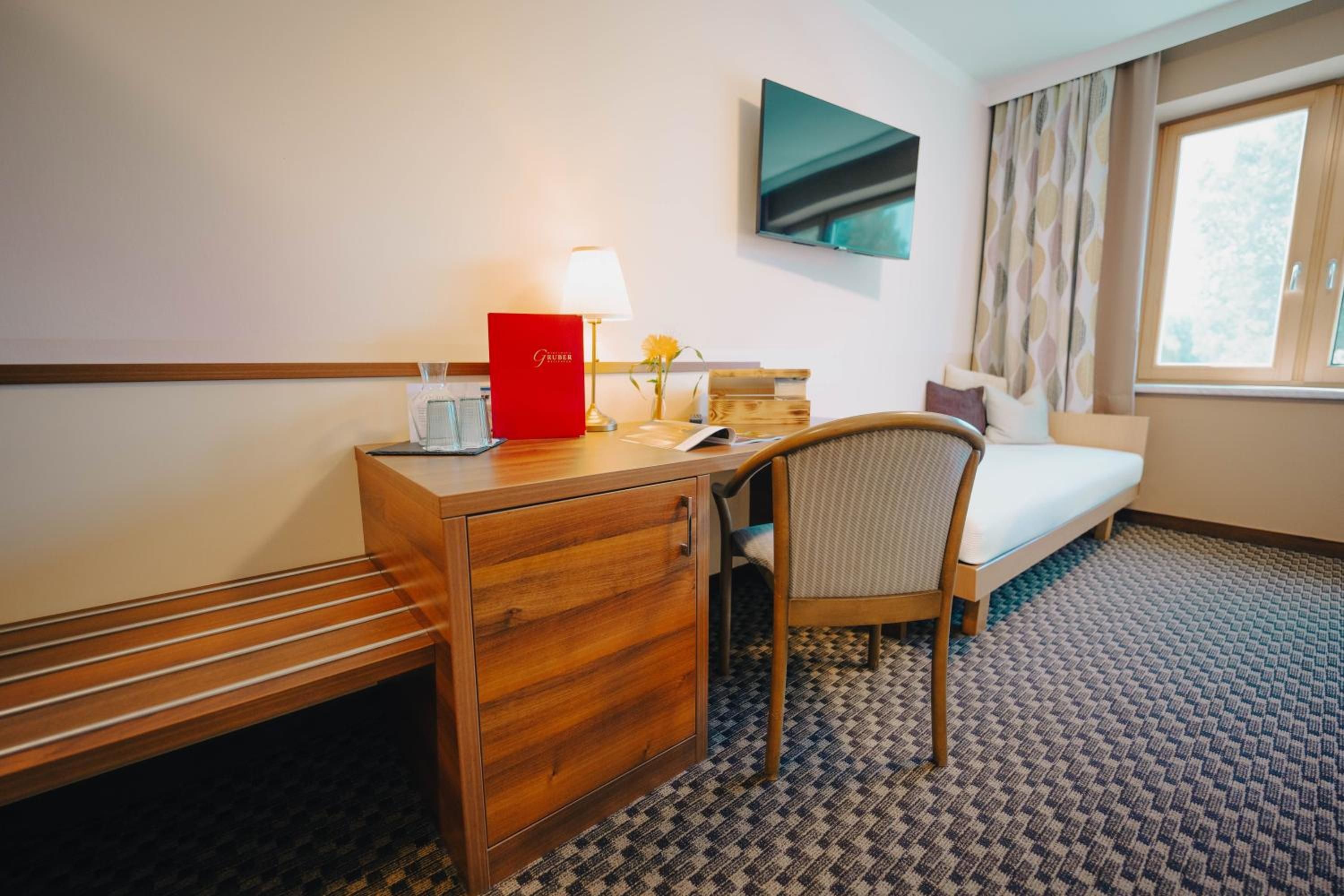 A hotel room with a wooden desk, a chair, a bed, a TV mounted on the wall, and a window with curtains.