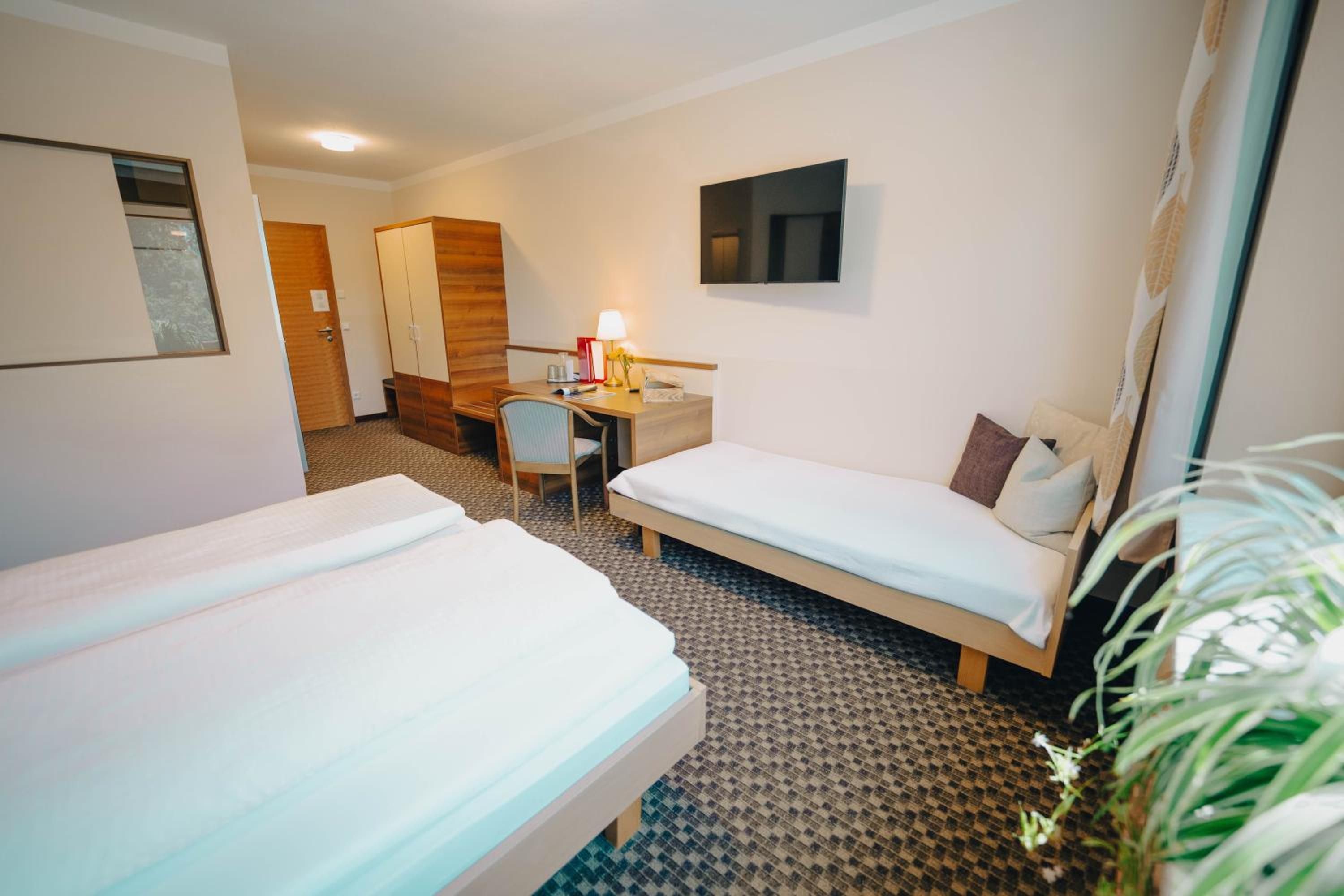A hotel room with two single beds, a desk with a chair, a TV mounted on the wall, and a small window.