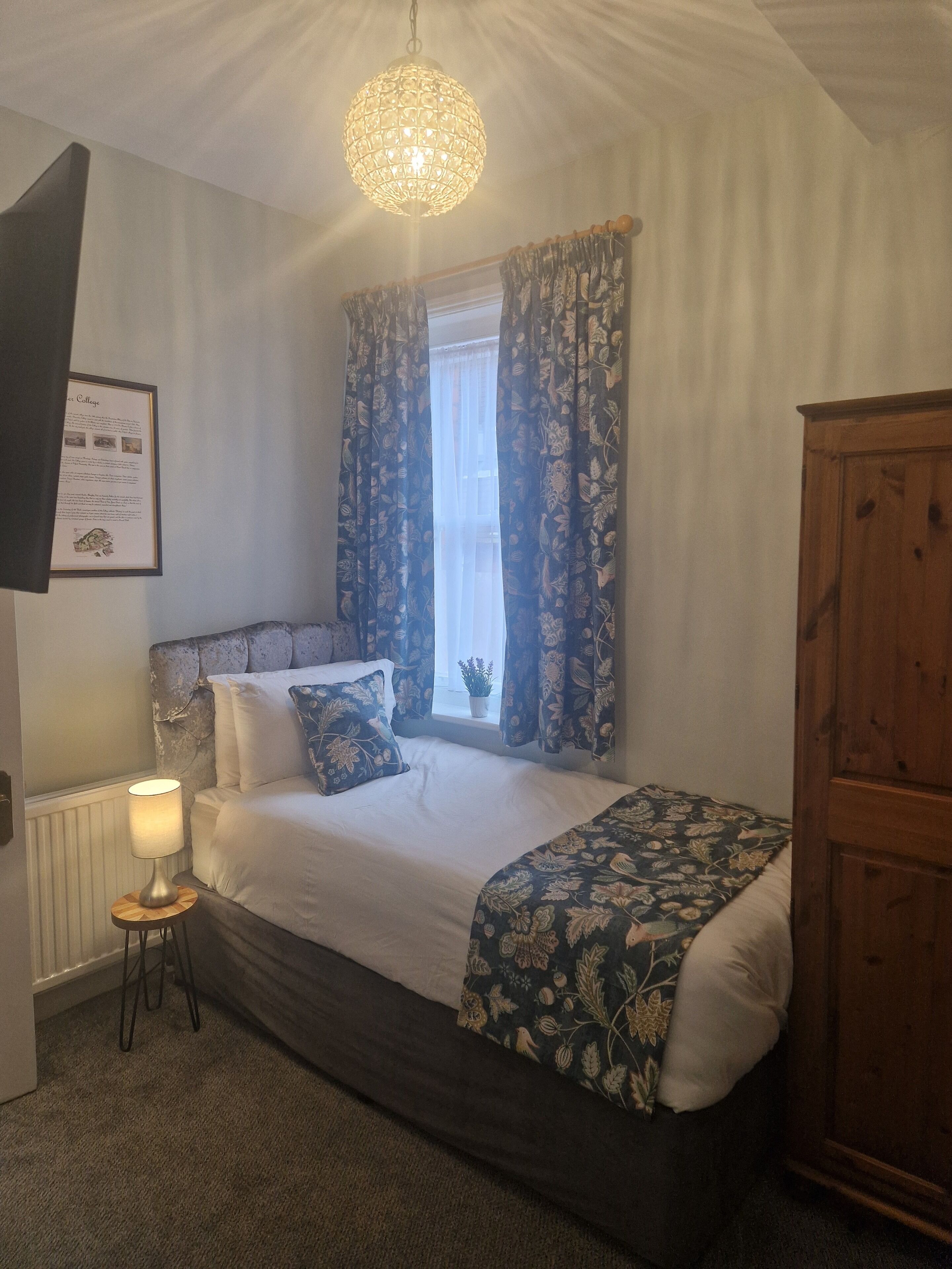 Quarto com cama, mesa de cabeceira, luminária, guarda-roupa de madeira e um certificado emoldurado na parede.