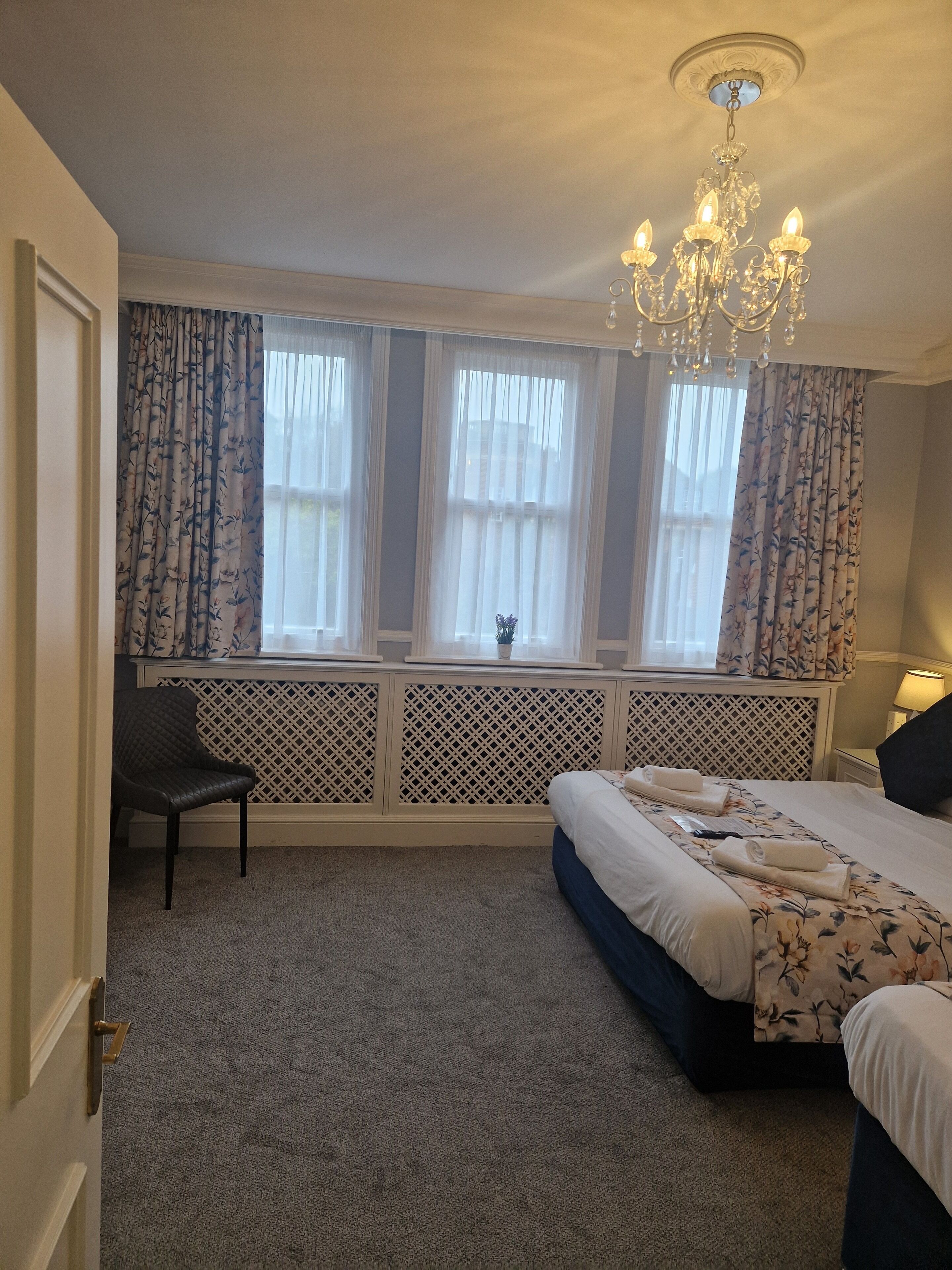 A bedroom with a large bed, a chair, a window with curtains, and a chandelier.