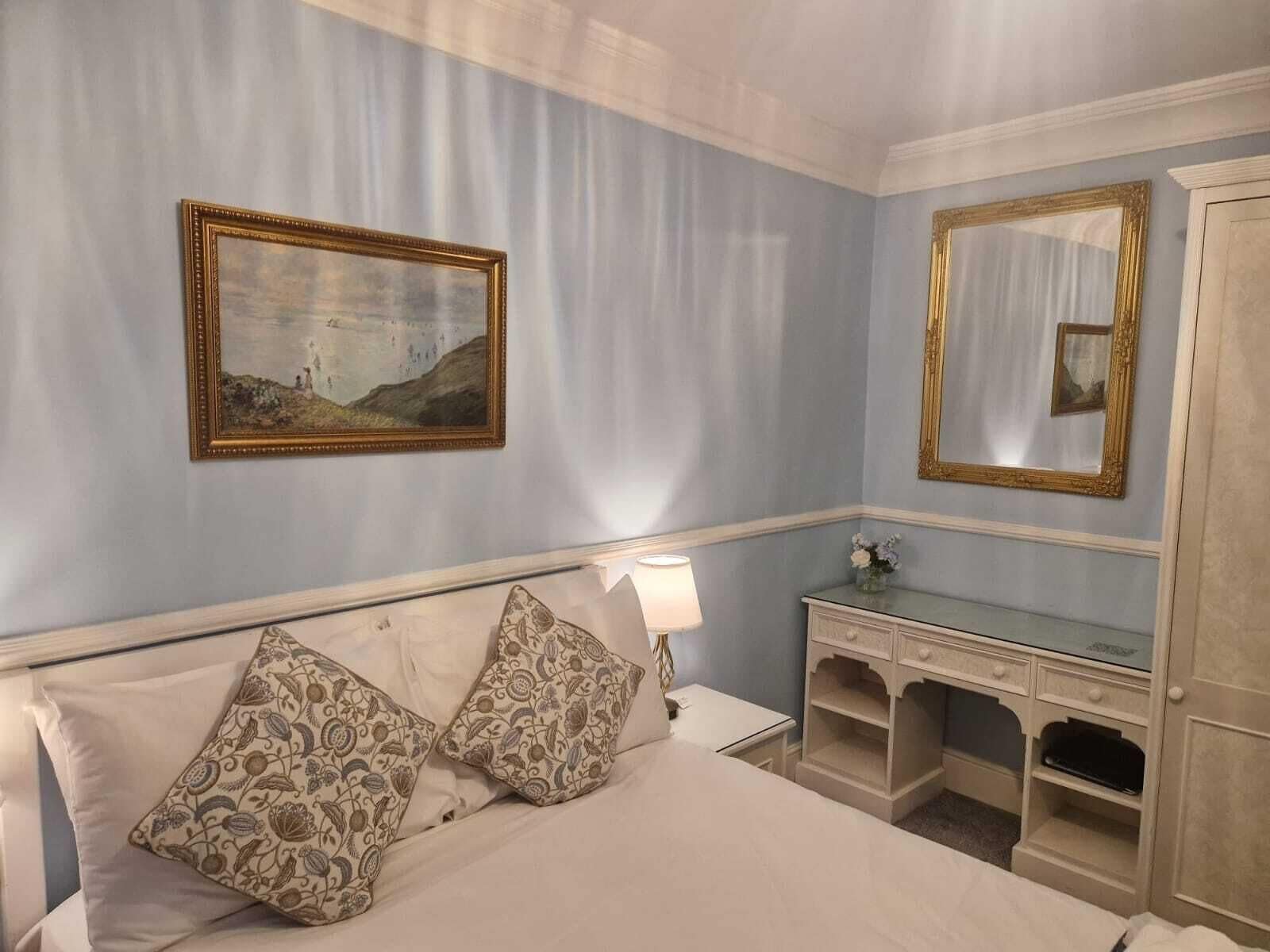 A hotel room with a bed, a nightstand, a desk, a mirror, and a painting on the wall.