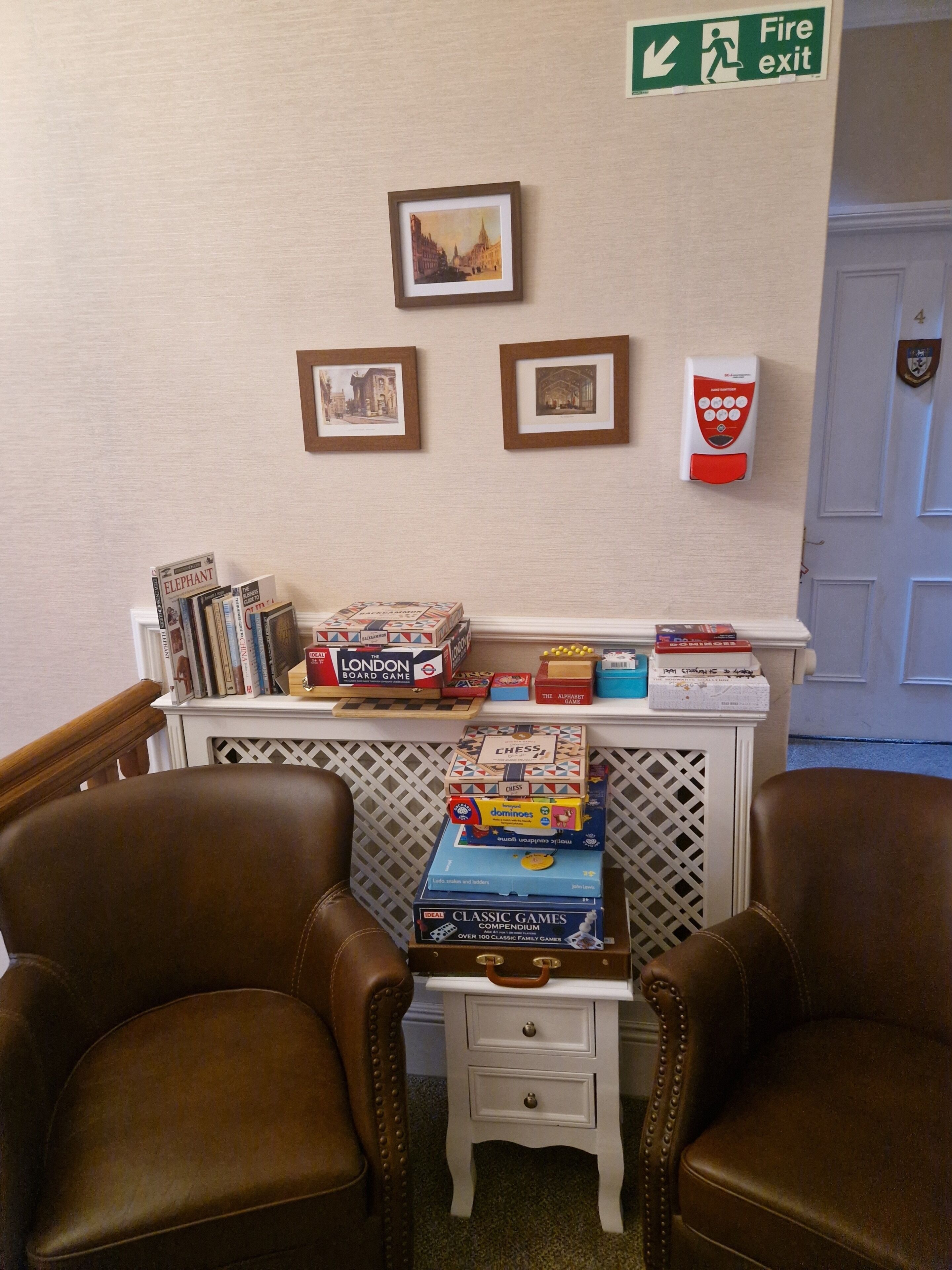 Ein Zimmer mit zwei braunen Ledersesseln, einem kleinen weißen Tisch mit Brettspielen, gerahmten Bildern an der Wand und einem Feuerschutzschild.