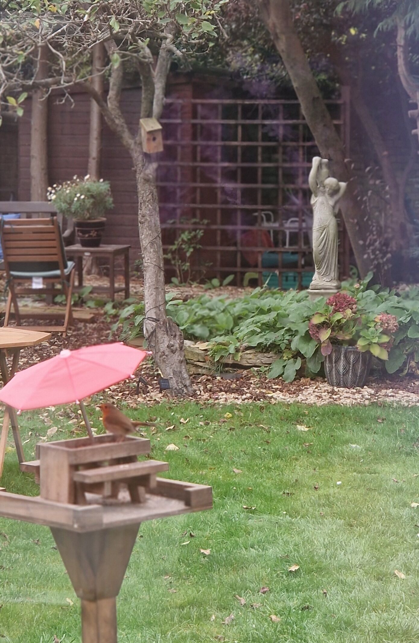 A garden with a wooden bird feeder, a statue, and a red umbrella.