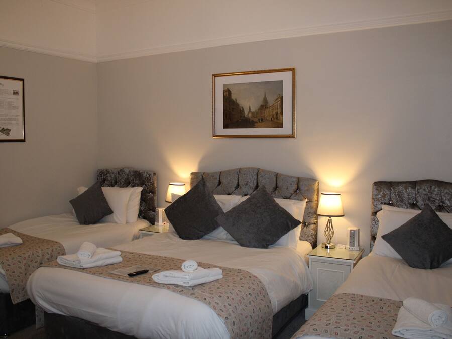 A hotel room with three beds, each with a headboard, pillows, and a bedside table with a lamp. There is a framed picture on the wall above the beds.
