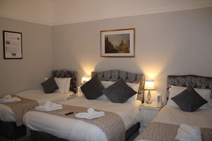Habitación de hotel con tres camas, cada una con cabecera, almohadas y mesita de noche con lámpara. Hay un cuadro enmarcado en la pared sobre las camas.