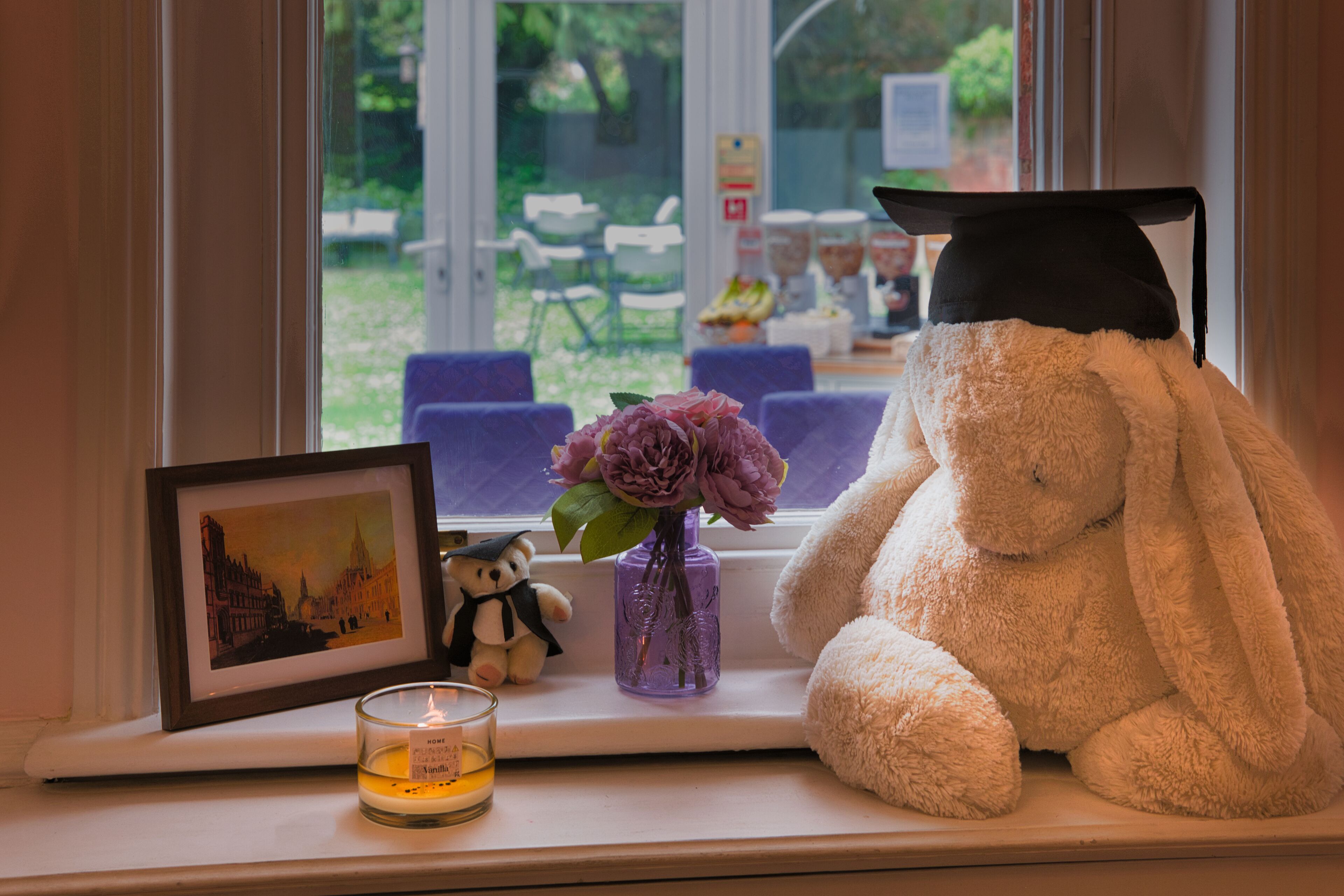 A window sill with a framed picture, a candle, a vase with flowers, and a stuffed bunny wearing a graduation cap.