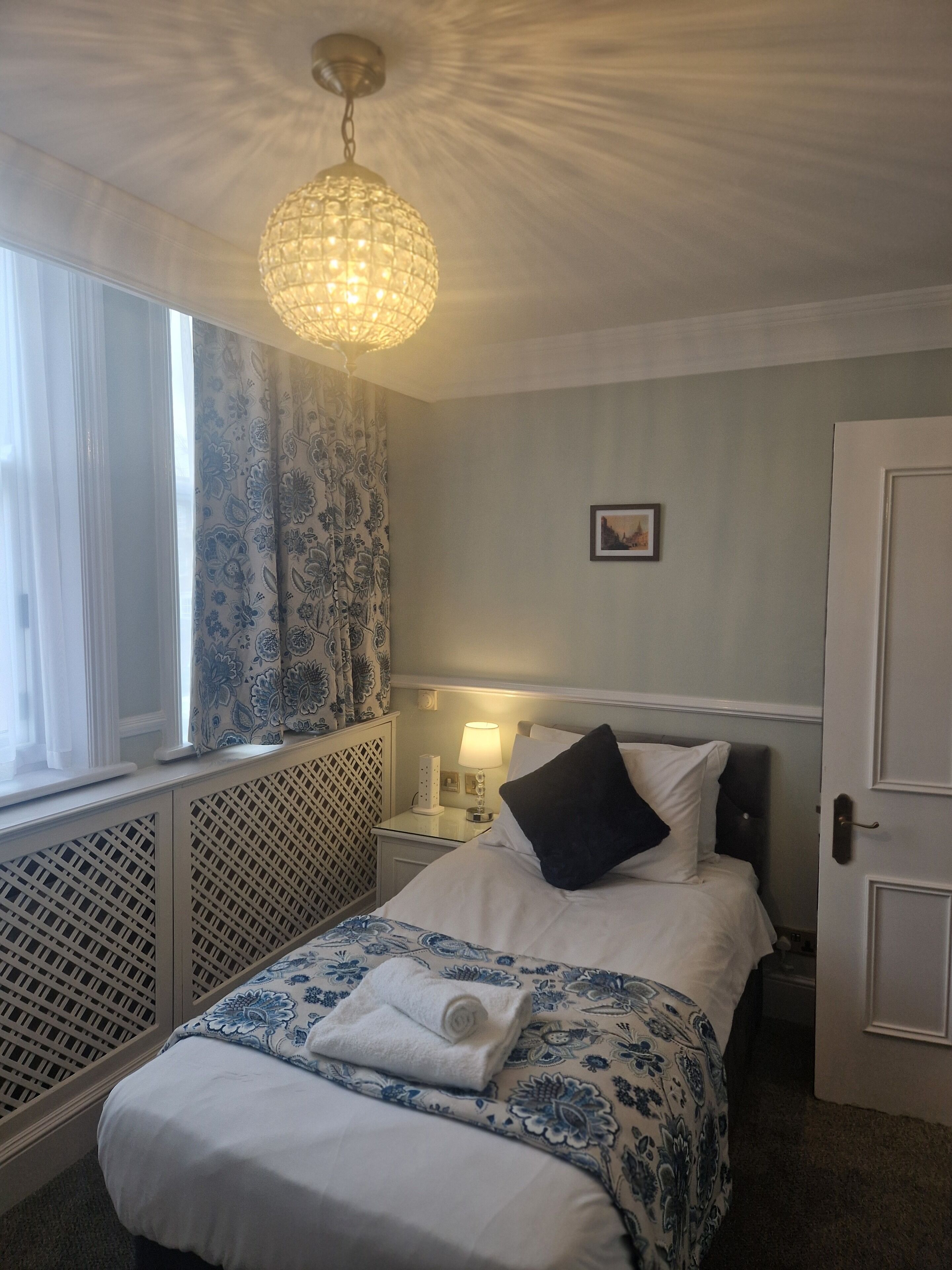 Una cama bien hecha con una colcha blanca y azul estampada con flores, una toalla blanca enrollada y una almohada negra. Se observa una mesita de noche con una lámpara, una ventana con cortinas azules estampadas con flores y un cuadro colgado en la pared.