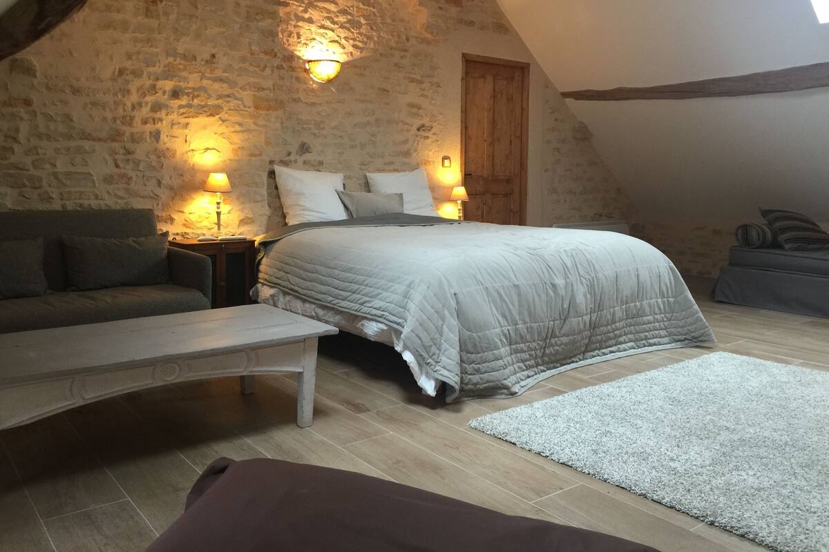 A cozy bedroom with a stone wall, a bed with a grey duvet, a wooden bench, and a small rug.
