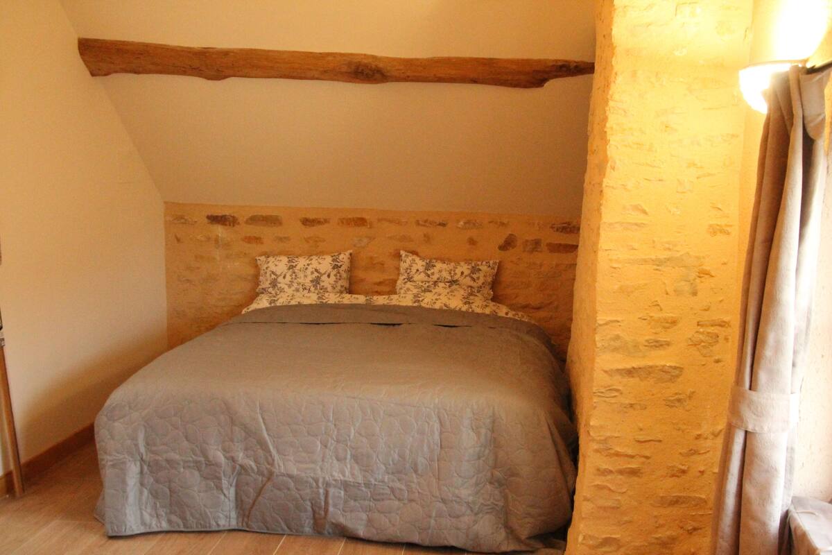 A bedroom with a stone headboard, a bed with a grey quilt, and a wooden beam ceiling.