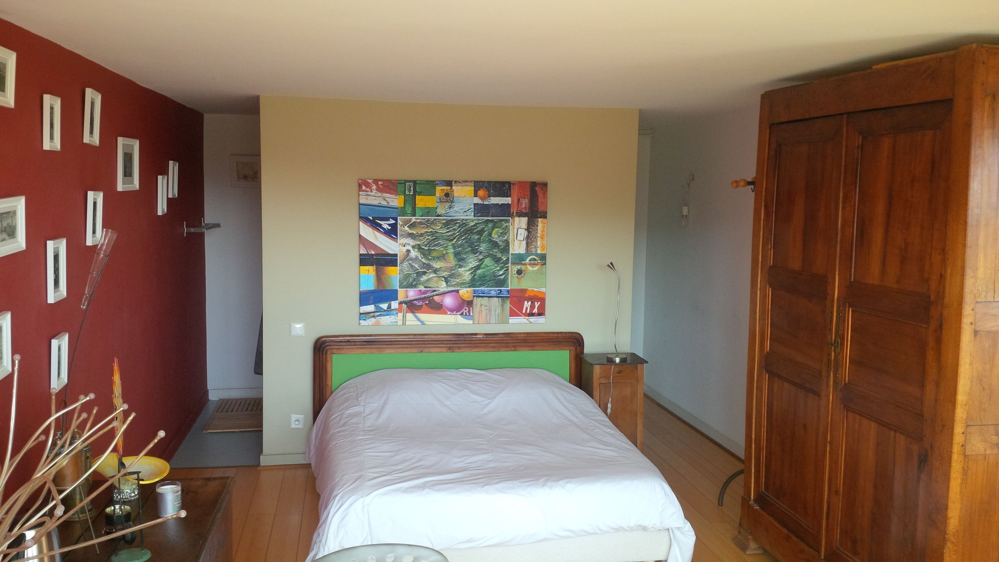 A bedroom with a bed, a wooden wardrobe, a wall with framed pictures, and a colorful abstract painting.