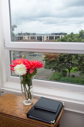 A window with a view of a building and trees, a vase with flowers, and a stack of books on a wooden table. - Beautiful City Center 2 Bed Apartment with Park View (Edinburgh)