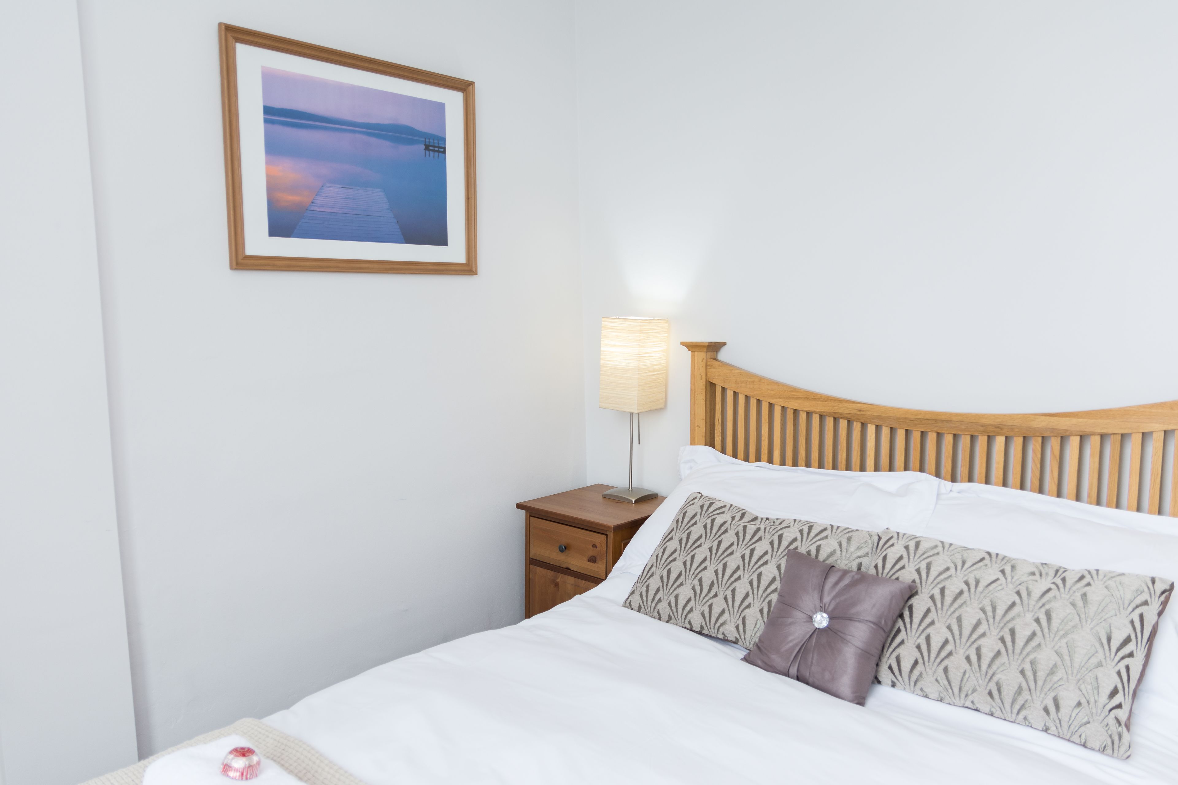 A neatly made bed with a wooden headboard, a bedside table with a lamp, and a framed picture on the wall.