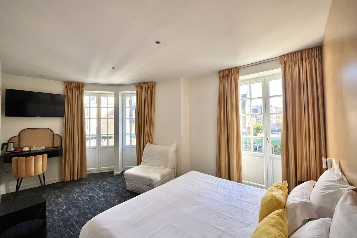 A hotel room with a bed, a desk, a chair, a TV, and large windows with curtains.