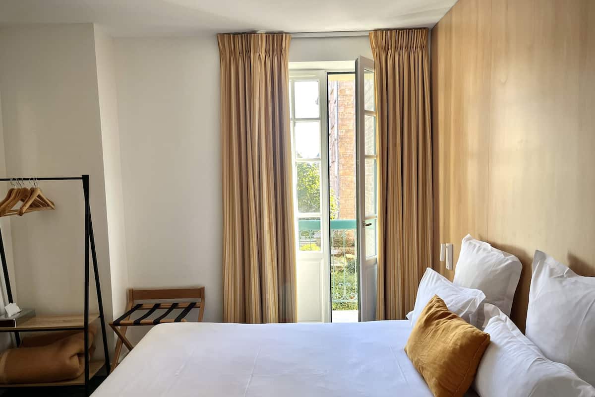 A hotel room with a bed, a wooden chair, a coat rack, and a view of the outside.