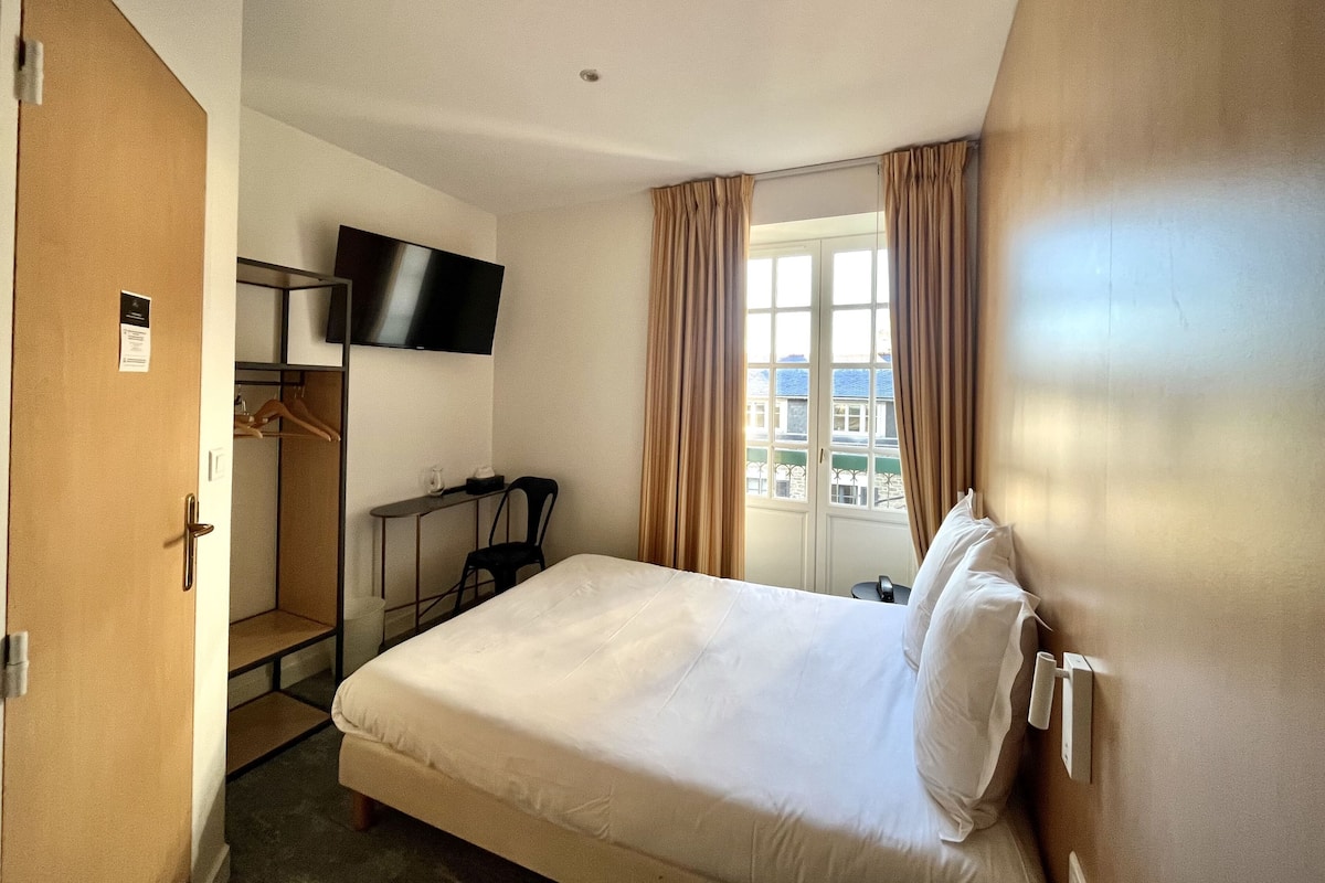 A hotel room with a bed, a desk, a chair, a TV mounted on the wall, and a window with curtains.