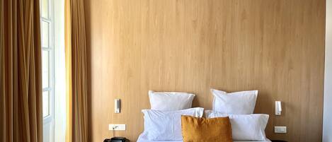 Quarto de hotel com cabeceira de madeira, roupa de cama branca, travesseiro cor mostarda e uma pequena mesa de cabeceira com telefone e carregador.
