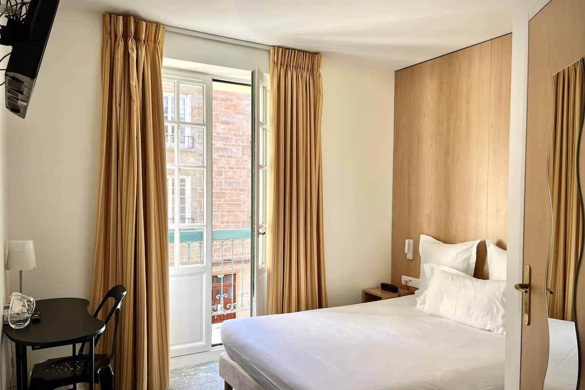 A hotel room with a bed, a desk with a chair, a telephone, and a view of a brick building through a glass door.