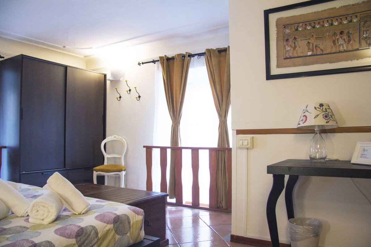 A hotel room with a bed, a chair, a table with a lamp, curtains, and a wall-mounted picture.
