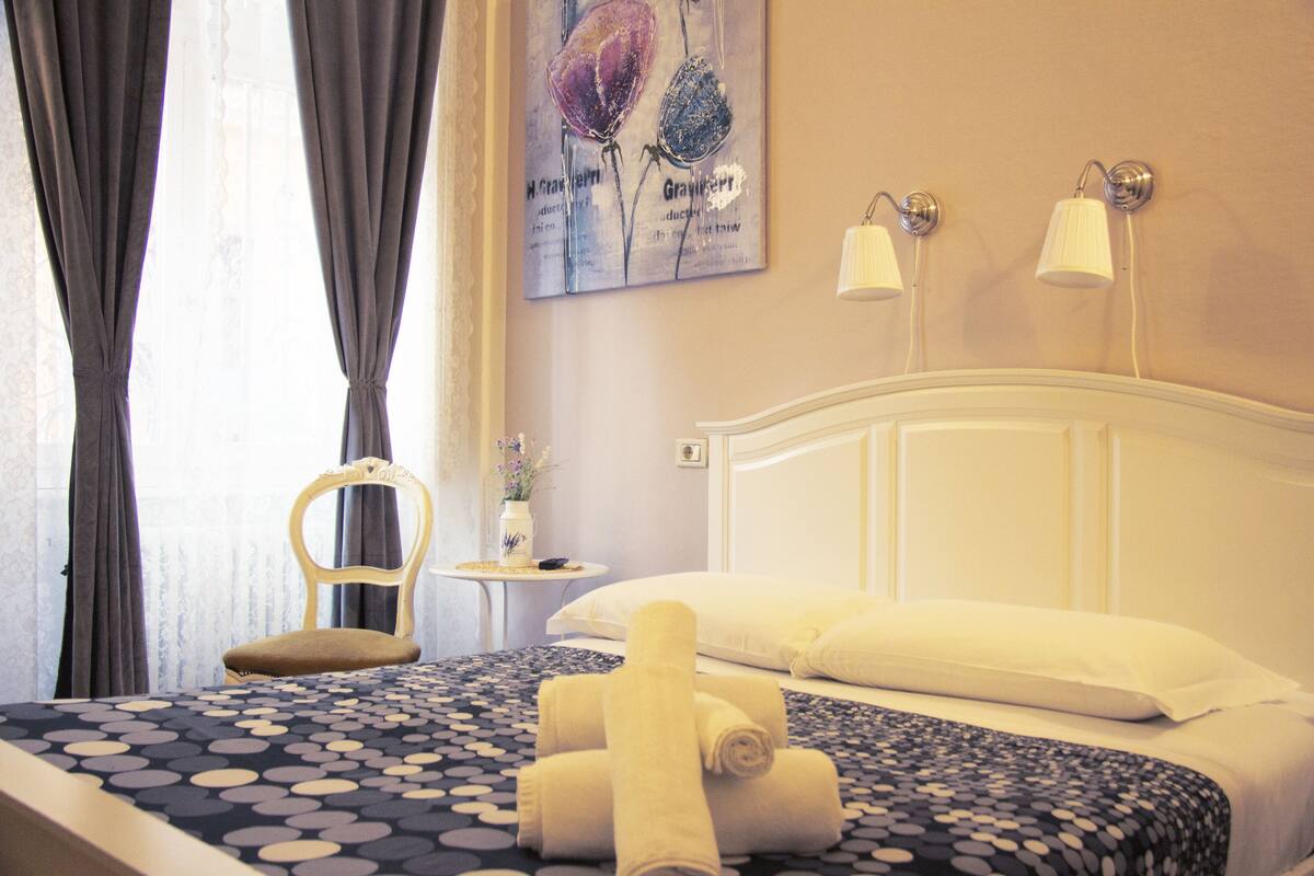 A hotel room with a bed, a chair, a small table with a vase, and a painting on the wall.