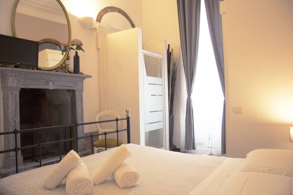 A hotel room with a bed, a fireplace, a mirror, and a window with curtains.