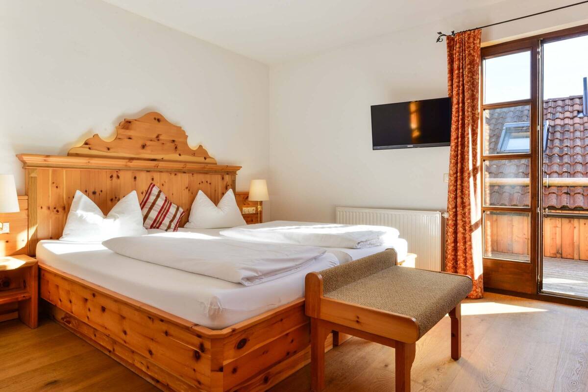 Ein hölzernes Bett mit weißen Leinentüchern, ein Nachttisch aus Holz mit einer Lampe, ein Fernseher an der Wand und eine Glastür mit Vorhängen.