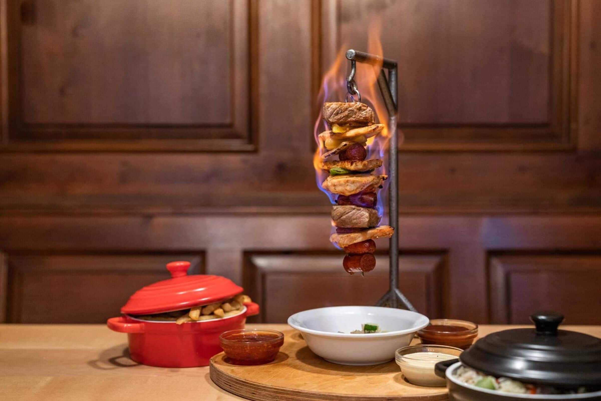 A kitchen with a meat skewer over a flame, a red pot, a white bowl, and various condiments on a wooden board.