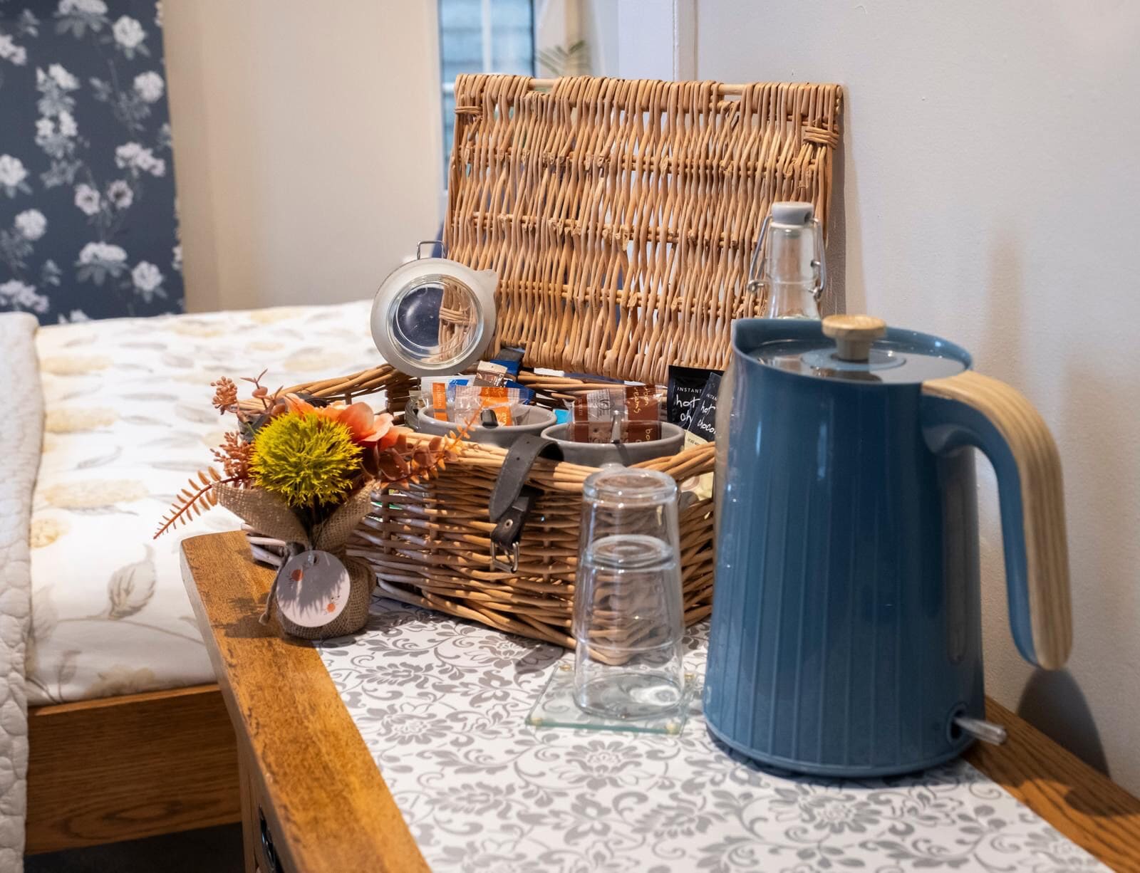 A wicker basket with a lid, a blue kettle, a glass jar, and a small plant on a patterned tablecloth.