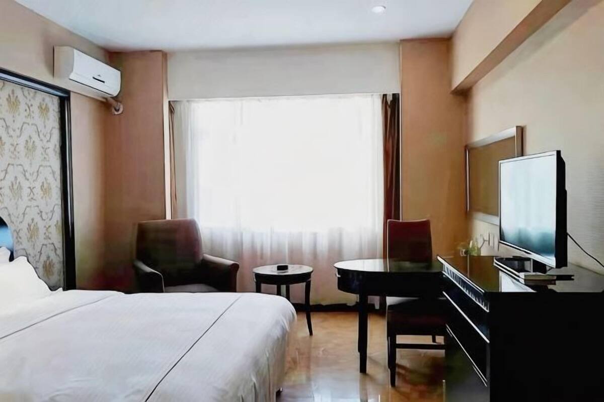 Ein Hotelzimmer mit Bett, zwei Stühlen, einem kleinen Tisch, einem Fernseher und einem Fenster mit Vorhängen.