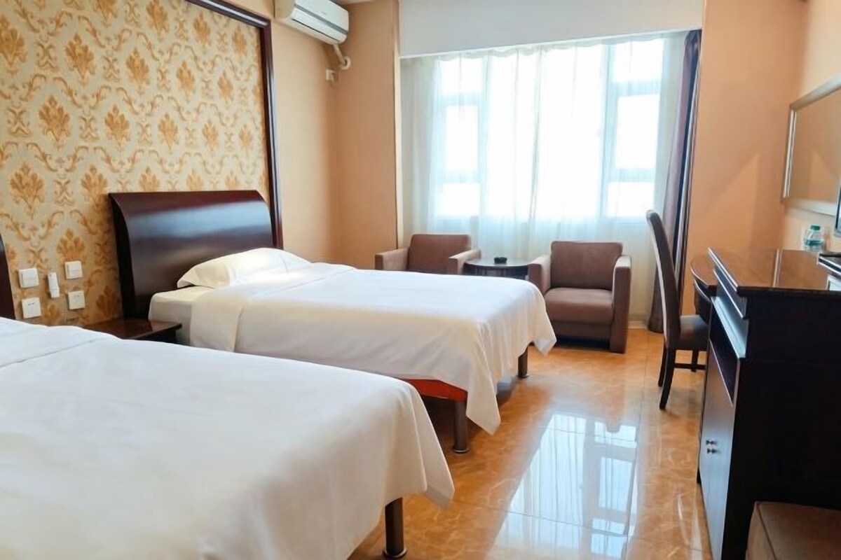 Quarto de hotel com duas camas, uma escrivaninha, uma cadeira e uma janela grande.