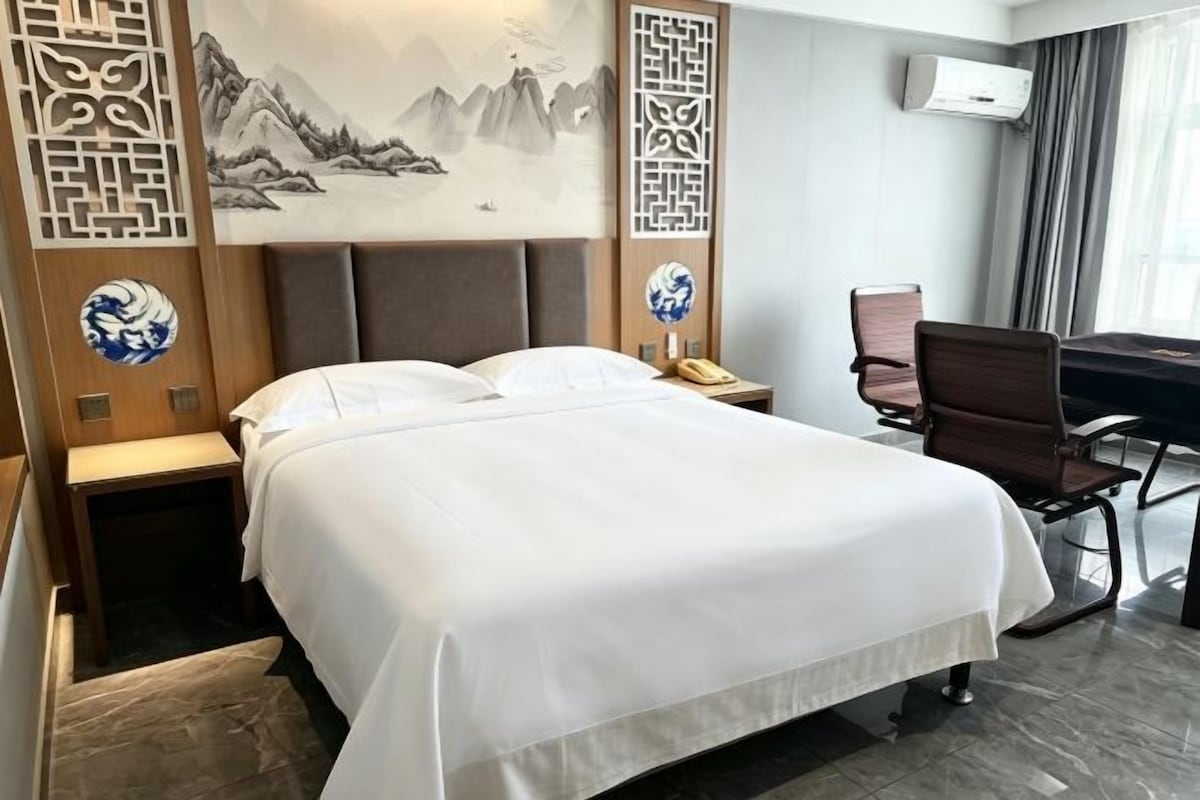 A hotel room with a bed, a desk, a chair, and a wall-mounted air conditioner.