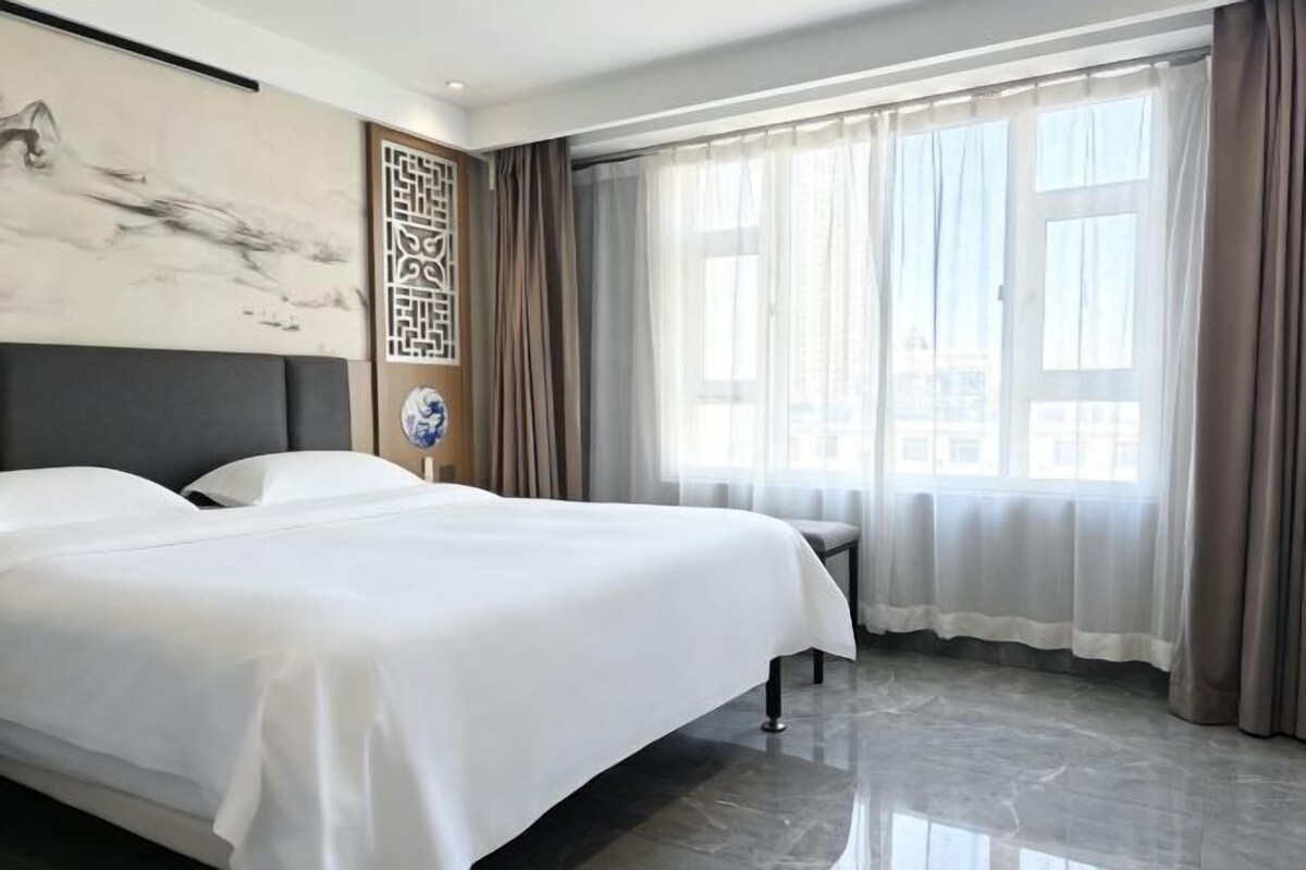 A hotel room with a large bed, a nightstand, a wall-mounted artwork, and a window with curtains.