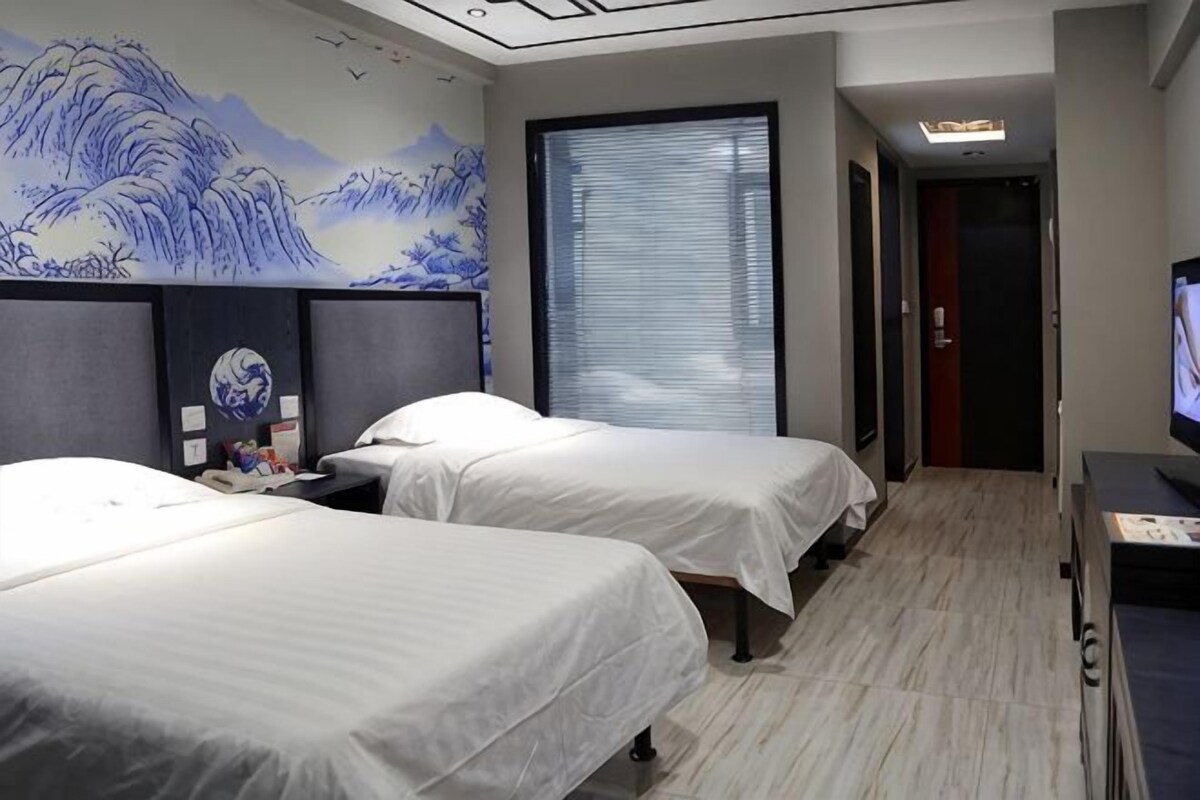 A hotel room with two beds, a large window with blinds, a television, and a decorative wall mural.