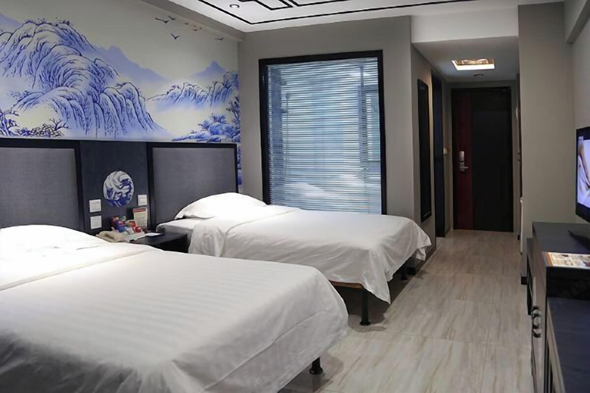 A hotel room with two beds, a large window with blinds, a wall mural, and a TV.