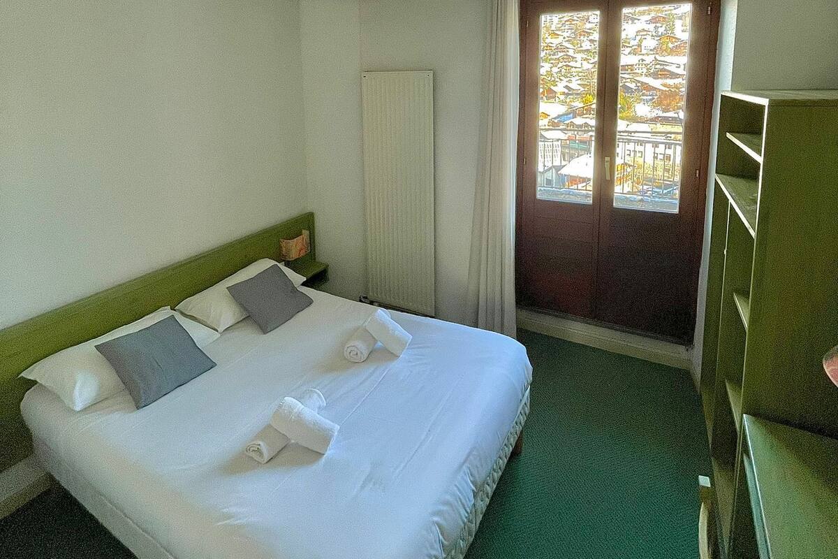 Une chambre avec un lit simple, une tête de lit verte, une table de chevet, un radiateur, une fenêtre avec des rideaux et une vue sur les bâtiments environnants.