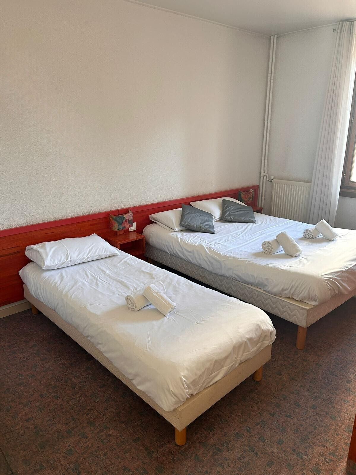 Two single beds with white bedding and rolled towels, a red headboard, and a window with curtains.