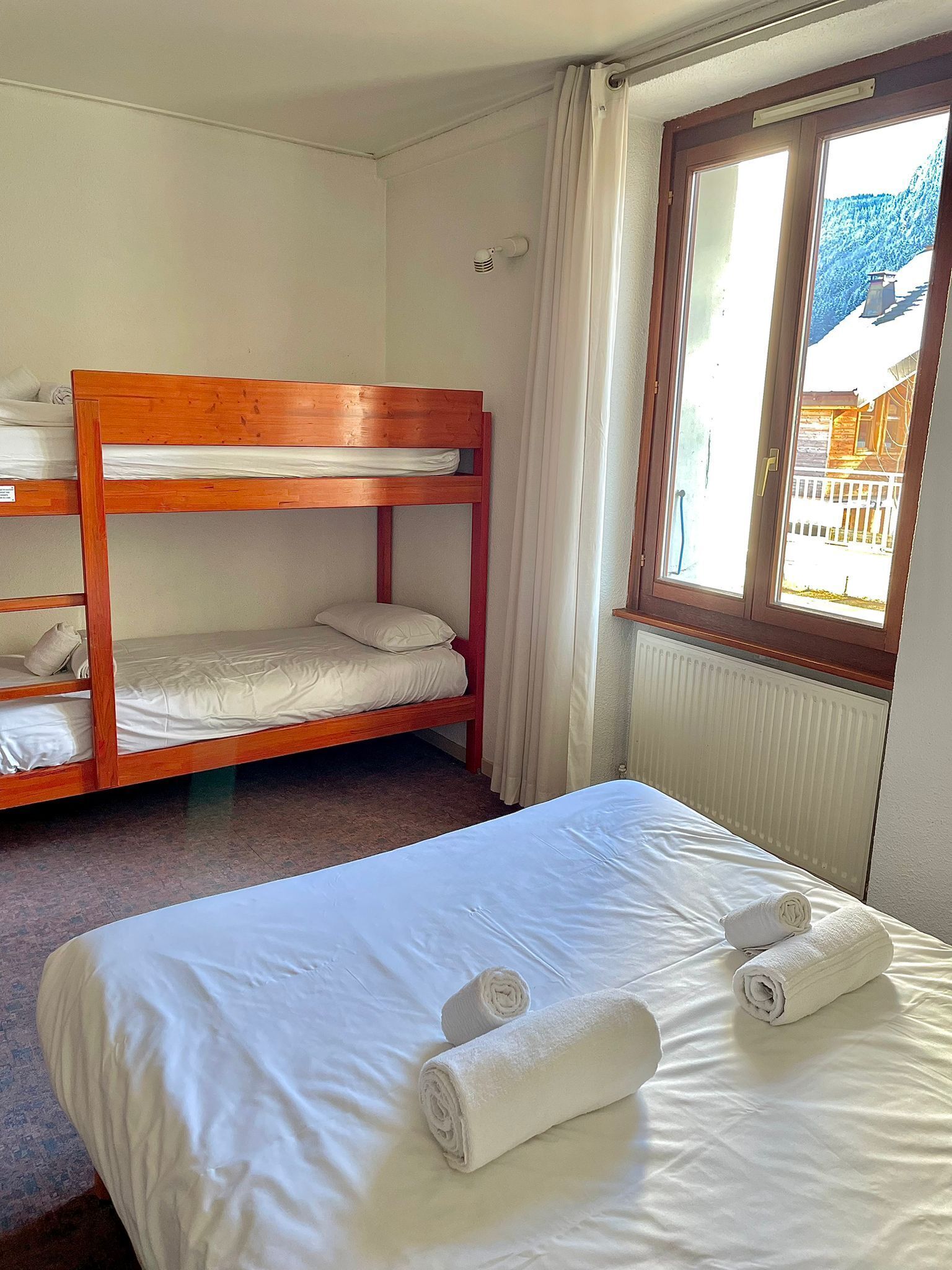 A room with a bunk bed, a single bed with rolled towels, a window with a view of a mountain, and a radiator.