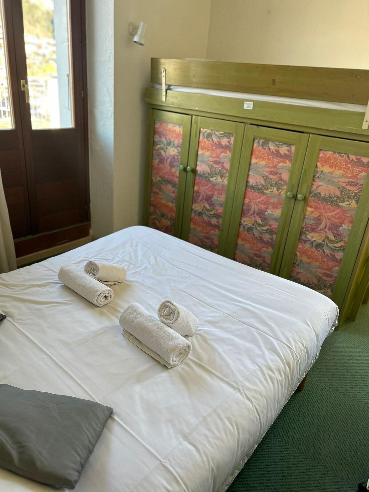 A neatly made bed with white linens and rolled towels, a green upholstered headboard with floral patterns, and a green carpeted floor.